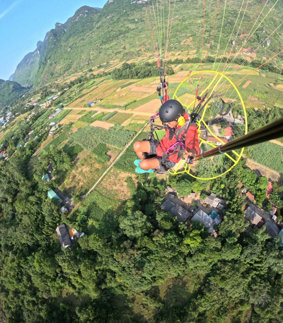 Phi công PPG bay đơn ở độ cao thấp trên ruộng và bản làng, sườn đá vôi dựng ngay phía sau.